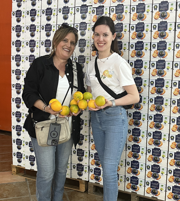 KI generiert: Zwei Frauen halten Orangen vor gestapelten Kisten mit Orangensaftaufdruck.