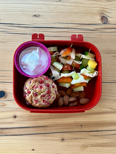 KI generiert: Eine Lunchbox mit Joghurt, Obstspießen, einem Muffin und Mandeln.