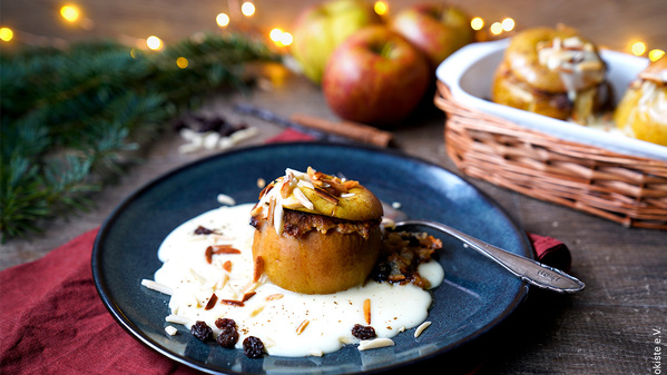 KI generiert: Das Bild zeigt einen Bratapfel, der auf einem Teller mit Vanillesoße und Rosinen serviert wird. Im Hintergrund sind weitere Äpfel und weihnachtliche Dekorationen zu sehen.