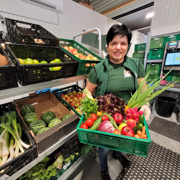 KI generiert: Eine Frau hält eine Kiste mit frischem Obst und Gemüse in einem Lagerraum.